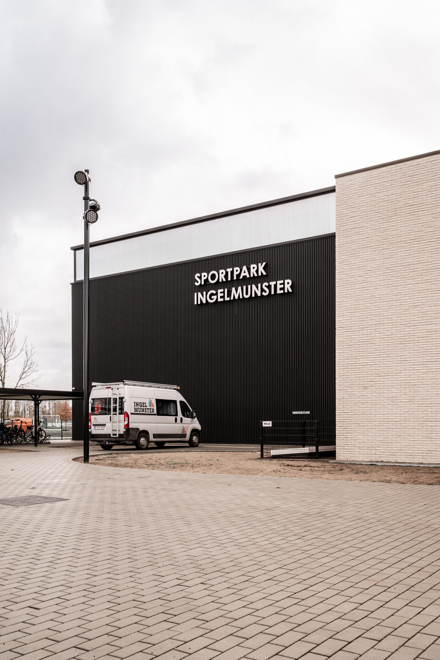 Vooraanzicht van Sportpark Ingelmunster door GC Architecten: een hoog volume met zwarte verticaal geribde gevelbekleding en witte letters, bereikbaar via een bestrate toegangsweg in halfsteens verband, geflankeerd door bestaande bomen en een overdekte fietsenstalling.