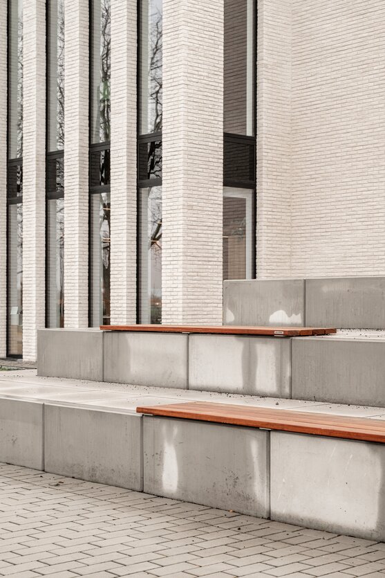 Zijaanzicht van Sportpark Ingelmunster door GC Architecten: een compact volume in lichte gevelsteen met slanke, hoge raampartijen, aansluitend op de zwarte sporthal, gezien vanaf het ruime bestrate voorplein.
