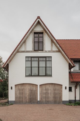 Voorgevel van een cottagevilla in Knokke door GC Architecten: een witgeschilderde bakstenen gevel met puntgevel, houten balken in de topgevel, twee rondbogige houten garagepoorten en donkere raampartijen met fijne roedeverdeling. | © Bert Demasure