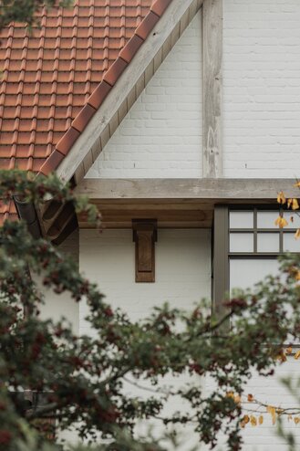 Detailzicht van een cottagevilla in Knokke door GC Architecten: houten balk en geprofileerde console onder de dakoverstek van een puntgevel, met rode pannen, witgeschilderde bakstenen gevel en een raampartij met fijne roedeverdeling, gezien door een waas van bladeren. | © Bert Demasure