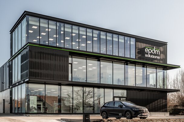 Straatzicht van het kantoor van EPDM Solutions in Roeselare door GC Architecten: een driedelig bedrijfsgebouw met gestapelde volumes in zwarte verticaal geribde gevelbekleding en grote glaspartijen, geaccentueerd door een groene lijn en het bedrijfslogo op de bovenste verdieping.