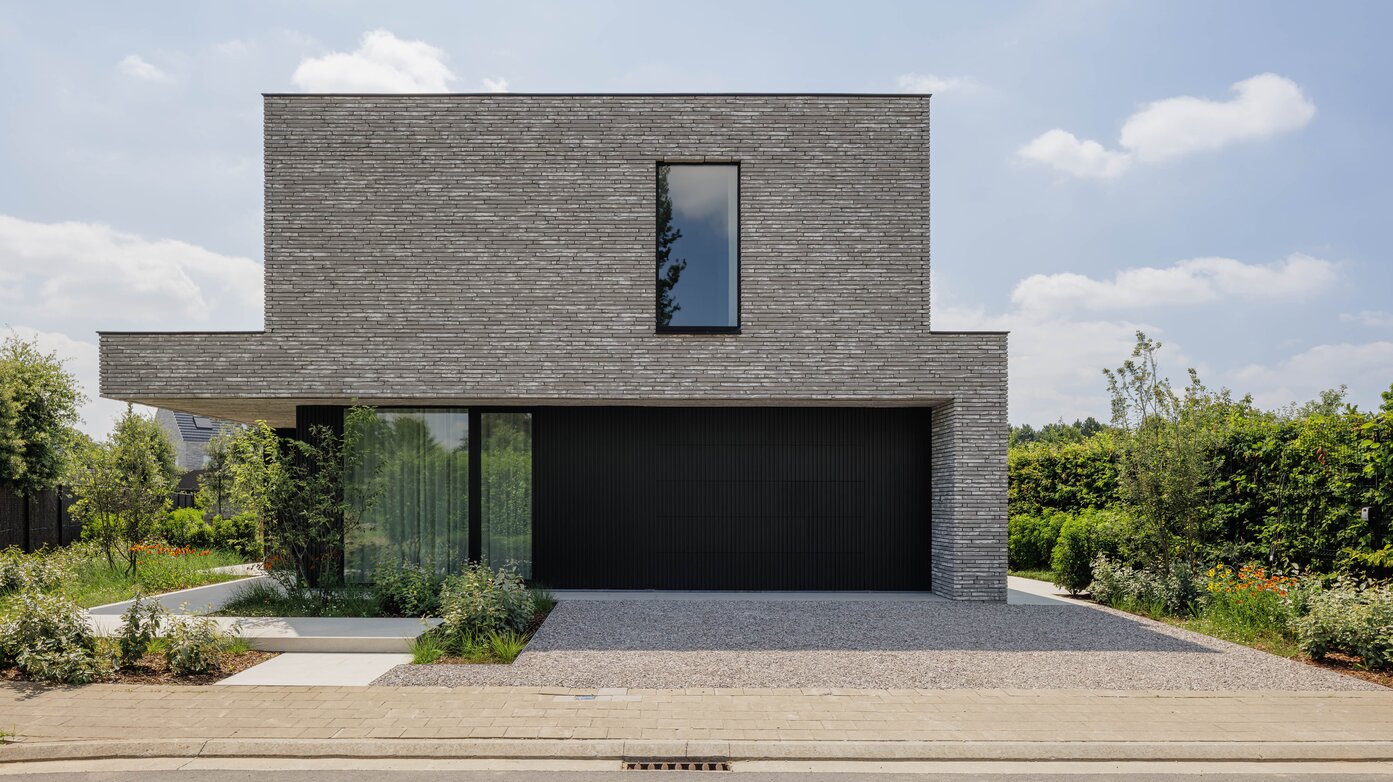 Straatzicht op een kubistische woning met grijze steenstrips, overkragend bovenvolume boven een zwarte garagepoort en een oprit met grind.