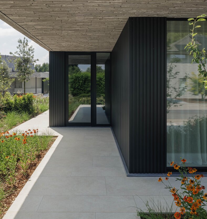 Overdekte wandelgang tussen twee gebouwvolumes met zwarte verticale lamellen, glazen deur en een pad van grote grijze terrastegels langs een bloemenborder.