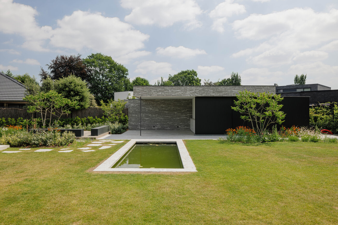 Laag tuinpaviljoen met grijze natuursteen gevel en zwarte lamellen, met rechthoekige vijver en bloemenborders in een strakke tuin, ontworpen door GC architecten.
