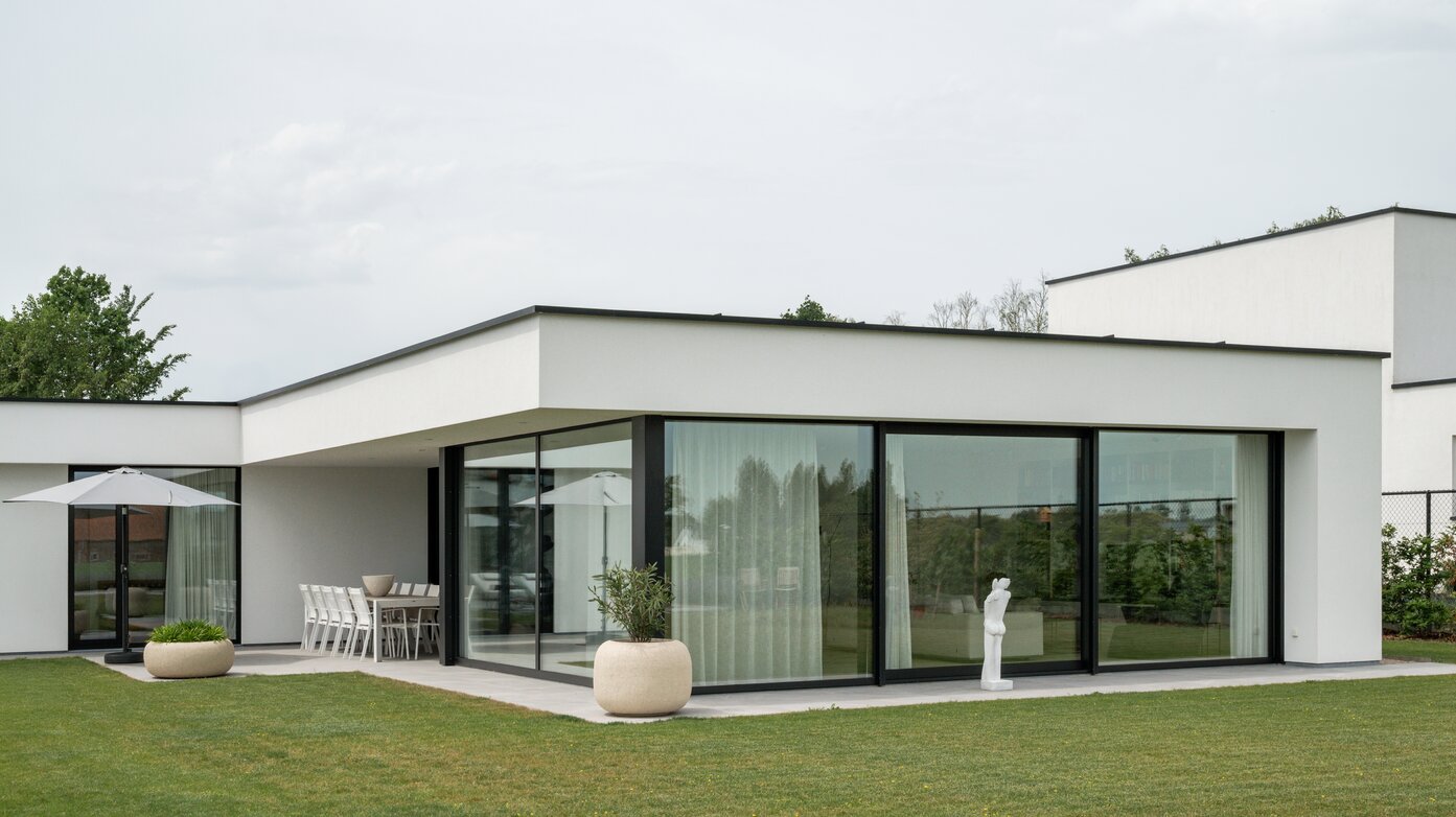 Wit gepleisterde villa met platte overkraging en glazen hoekraam, met overdekt terras, eettafel, parasol en een sculptuur op het gazon, ontworpen door GC architecten.