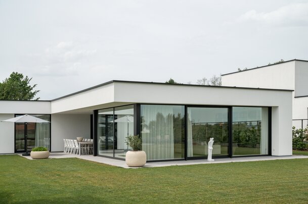 Wit gepleisterde villa met platte overkraging en glazen hoekraam, met overdekt terras, eettafel, parasol en een sculptuur op het gazon, ontworpen door GC architecten.