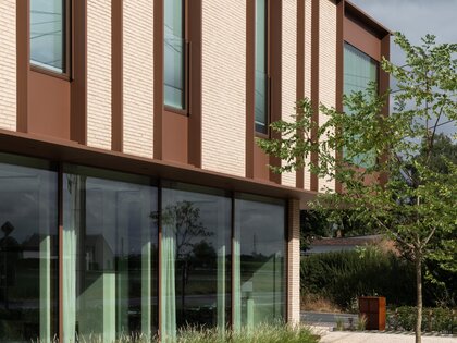 Diagonaal gevelaanzicht van een kantoorgebouw met crèmekleurige baksteen, verticale bronskleurige lamellen en een glazen pui op het gelijkvloers, ontworpen door GC architecten.