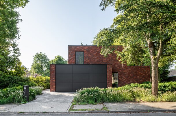 Voorgevel van een hedendaagse villa in donkerrode baksteen met een grote zwarte garagepoort, omgeven door groen en siergrassen.
