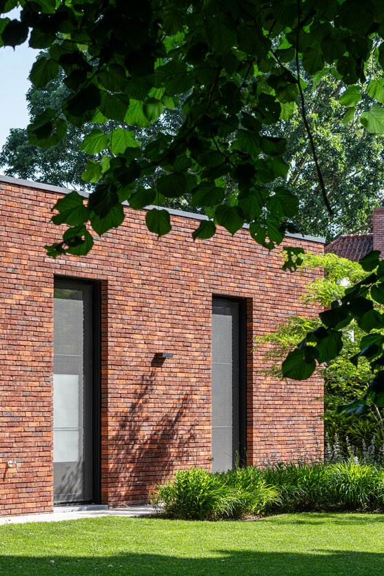 Bakstenen gevel met twee smalle, verticale raampartijen, gedeeltelijk overschaduwd door bladerdek.