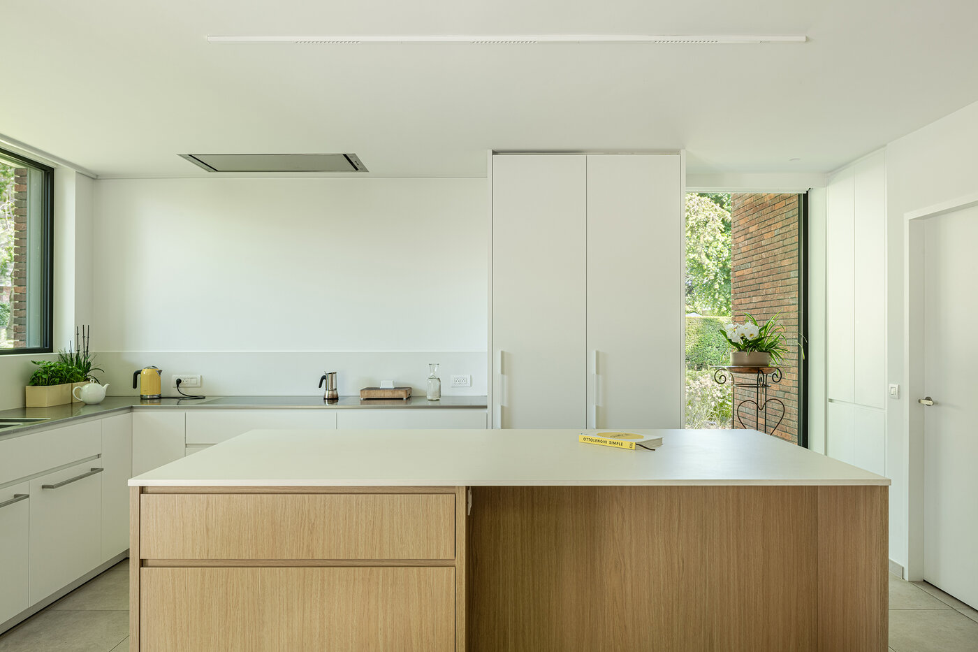 Lichte, minimalistische keuken met een houten kookeiland en wit meubilair, met zicht op de bakstenen buitengevel.