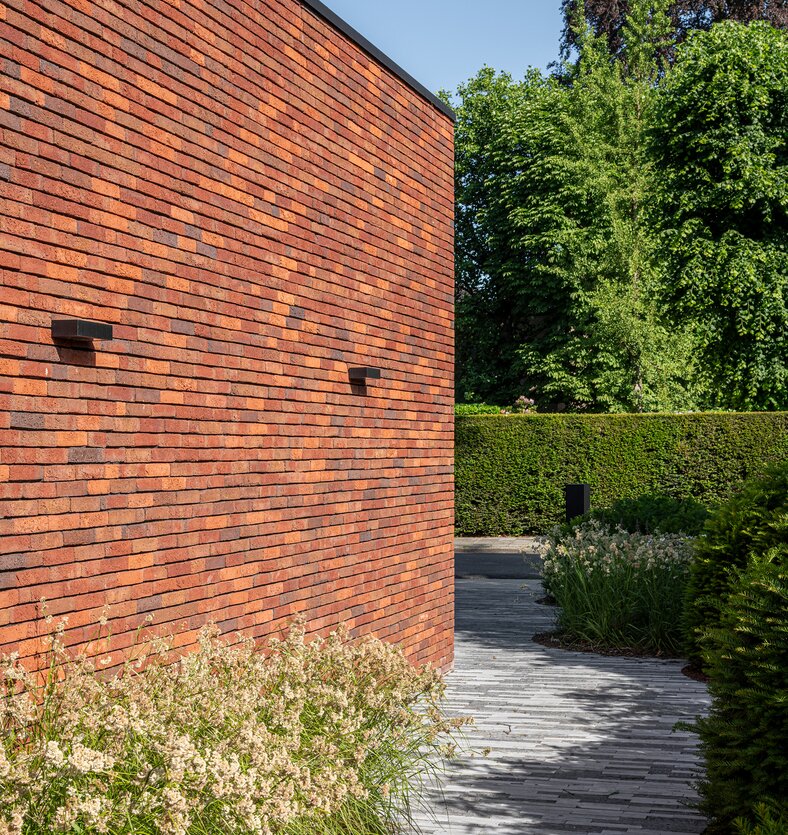 Close-up van een bakstenen gevelhoek met ingewerkte wandlampen, met een pad van grijze klinkers dat naar de tuin leidt.